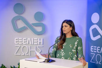A woman speaks at the ΕΞΕΛΙΞΗ ΖΩΗΣ podium holding a piece of paper in her hand